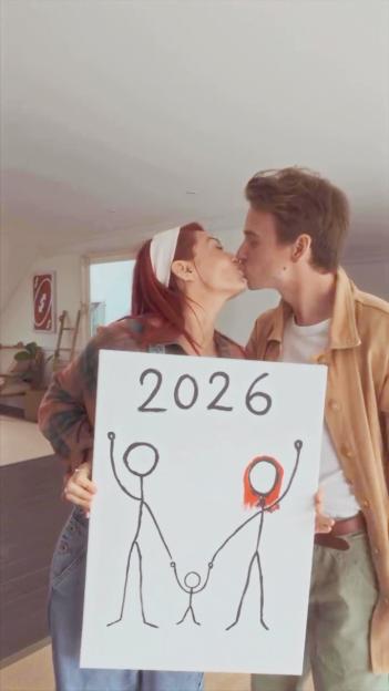 Dianne Buswell and Joe Sugg announcing their baby news with a sign showing stick figures of a family and the year 2026.