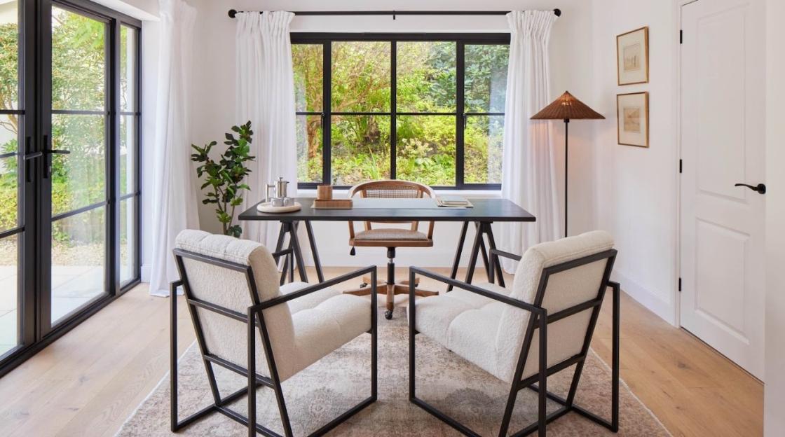 A home office with a dark desk, two white chairs, and large windows looking out to a lush garden.