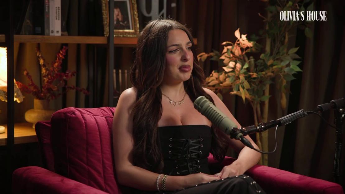 A young woman with long dark hair, wearing a black strapless top, sits in a red armchair, speaking into a microphone.