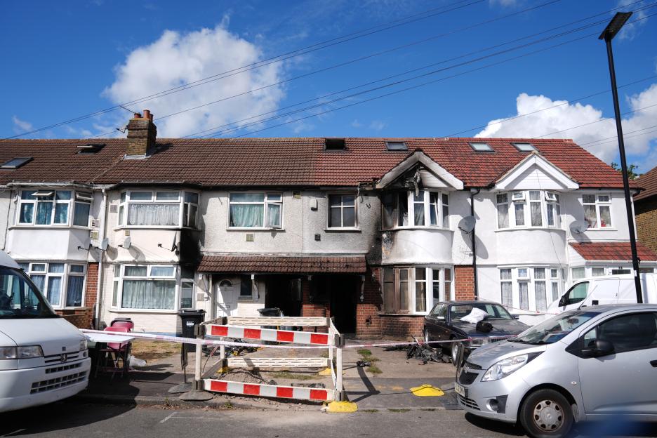 Terraced house with fire damage to one unit, cordoned off by police tape, with cars parked in front.