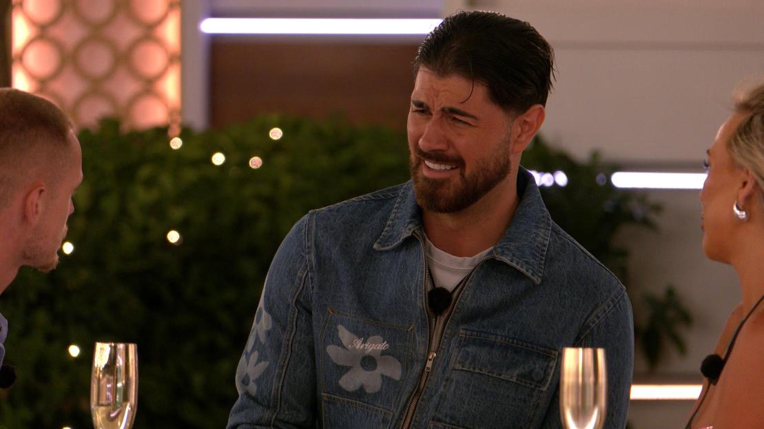 A man in a denim jacket with "Arigato" embroidered on it grimaces and looks to his right, while a woman and another man are partially visible on either side of him.