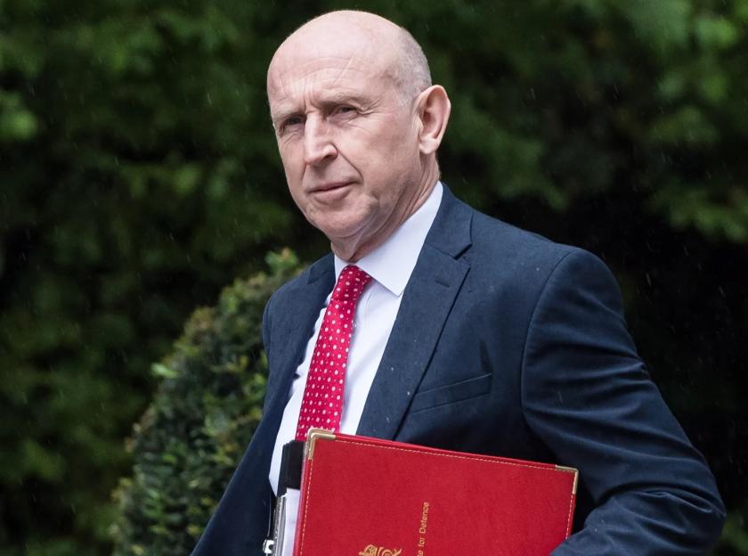 A man in a suit carrying a red binder labeled "Secretary of State for Defence" walks in the rain.