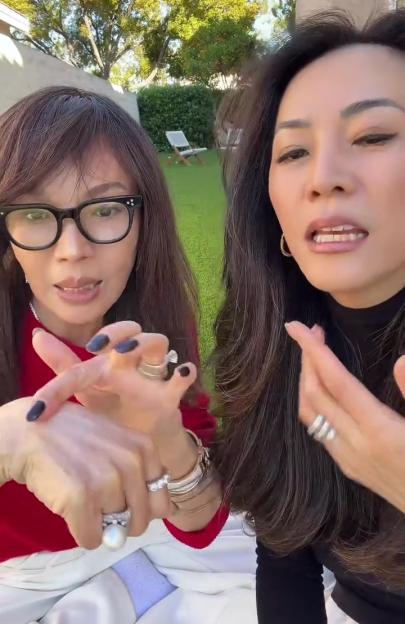 Two women with dark hair, one wearing glasses and a red top, and the other a black top, stand outdoors, gesturing with their hands.