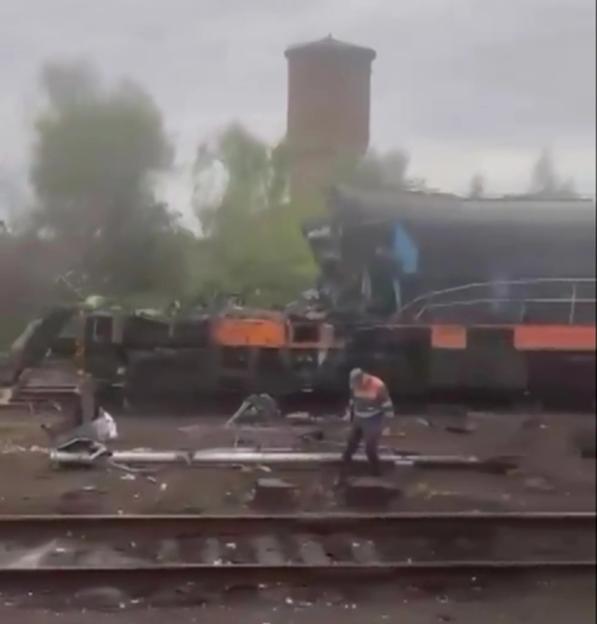 A damaged train with debris scattered on the tracks, and a person walking among the wreckage.