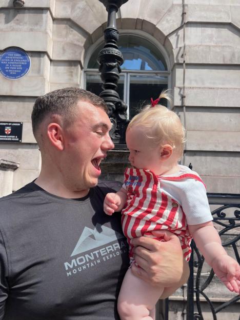 Jake Watts holding his daughter, Orla Ann, in front of a building with historical plaques.