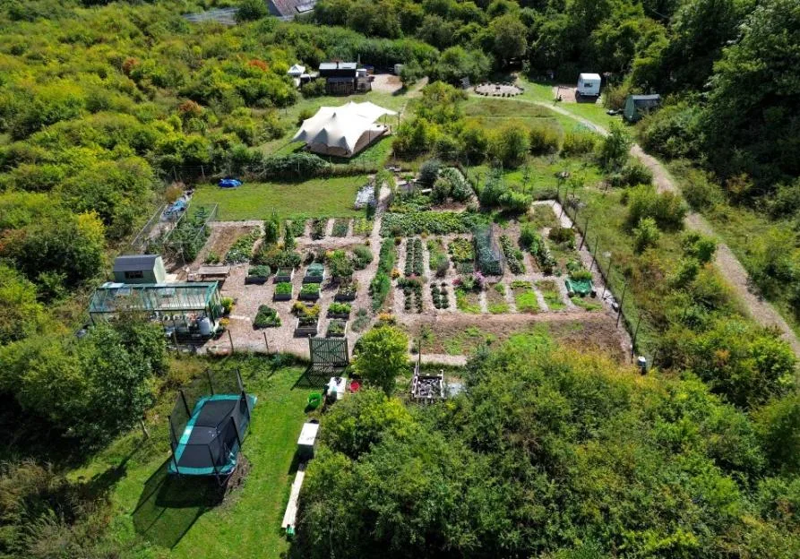 Aerial view of the off-grid Jolly Moon site in South Wonston, Hampshire, showing a large vegetable garden, greenhouse, trampoline, and tent structure surrounded by trees.