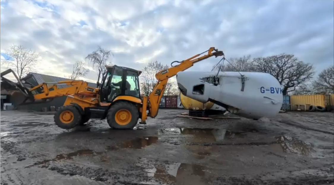 A yellow backhoe moving a partially dismantled white airplane fuselage in a muddy yard.