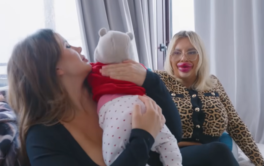 A woman with very large lips and glasses sitting next to another woman holding a baby.