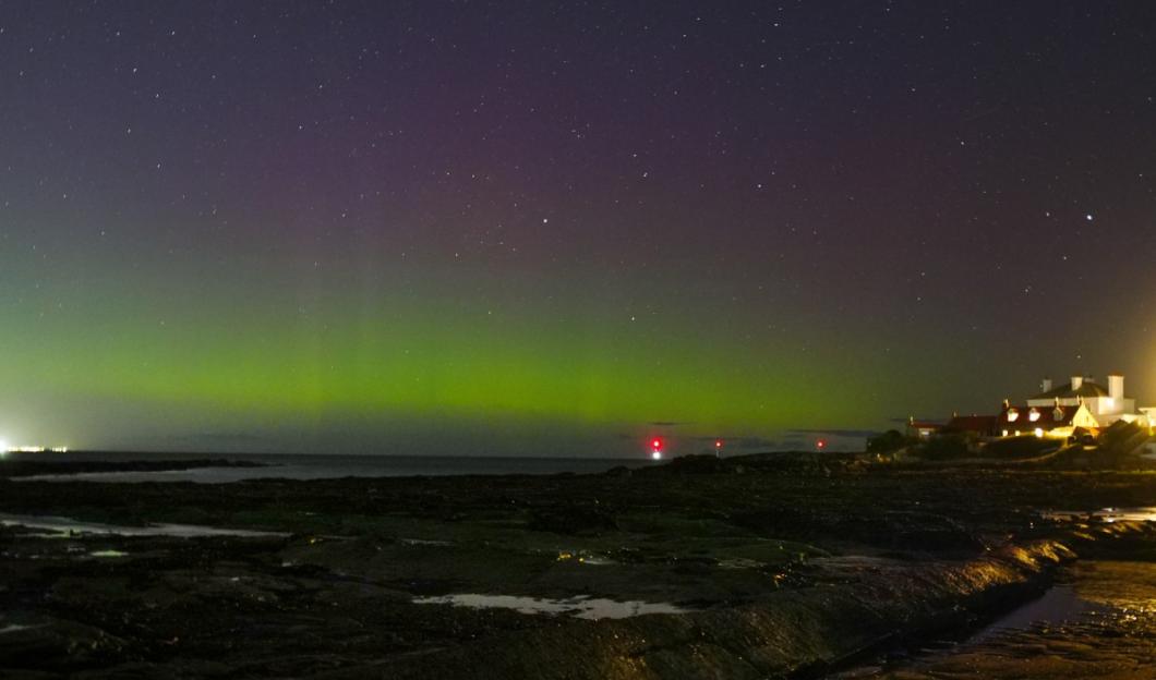 Northern lights over a rocky shore with distant houses and red lights.
