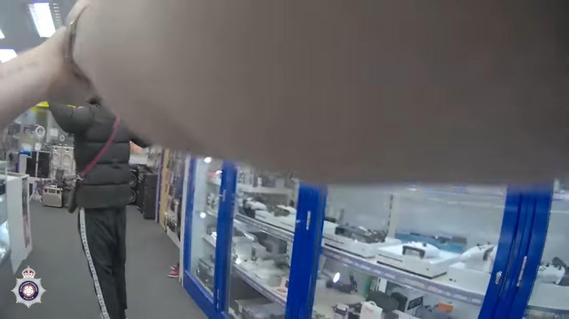 A robber, with hands raised, being arrested by a cop in a store filled with electronics.