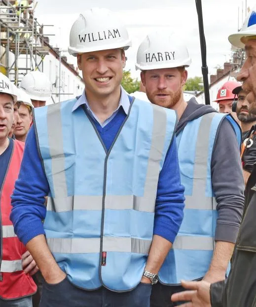 Princes William and Harry wearing hard hats labeled with their names and high-visibility vests at a DIY SOS building site.