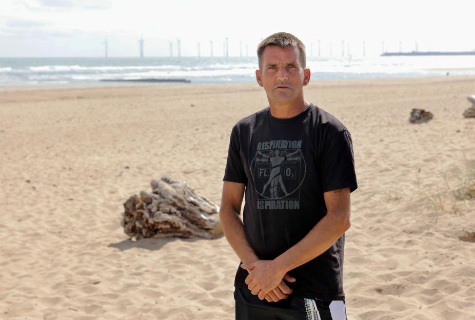 Davey Short, hero who saved a boy from drowning, stands on a sandy beach.