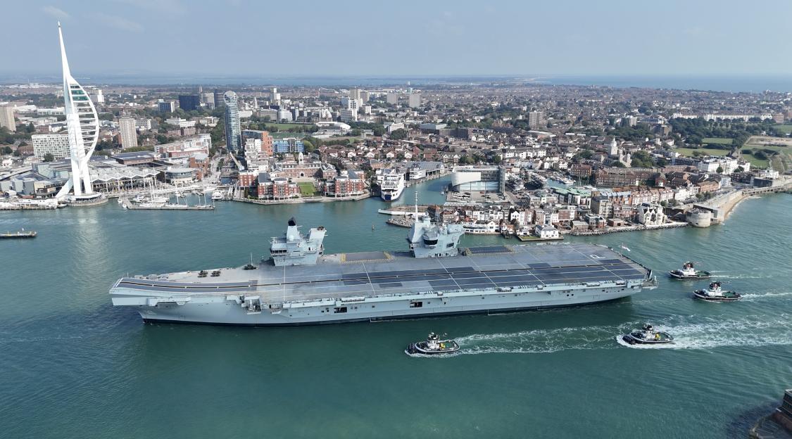 The HMS Prince of Wales aircraft carrier returns to Portsmouth Harbour, escorted by several tugboats, with the city and Spinnaker Tower in the background.