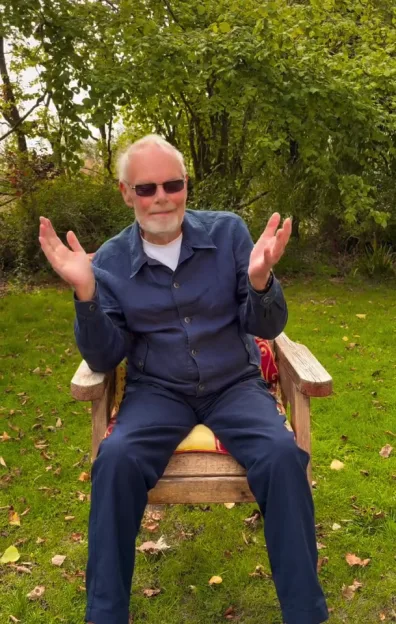 Bob Harris, also known as Whispering Bob, sitting in a wooden chair outdoors, wearing sunglasses and a navy blue jumpsuit, with trees and grass in the background.