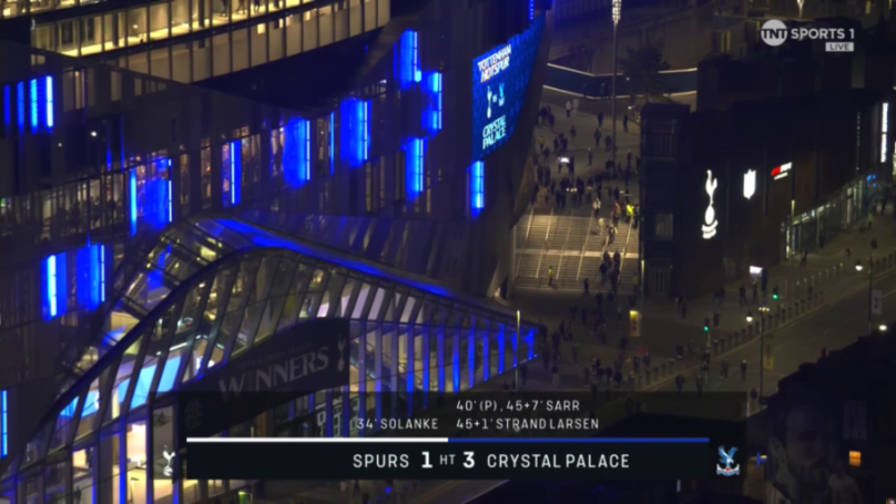 An aerial night view of a stadium building with blue lights and a scoreboard showing "Spurs 1 HT 3 Crystal Palace".