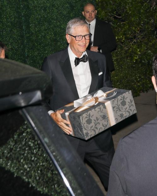 Bill Gates, smiling and wearing a tuxedo, holds a wrapped gift box.