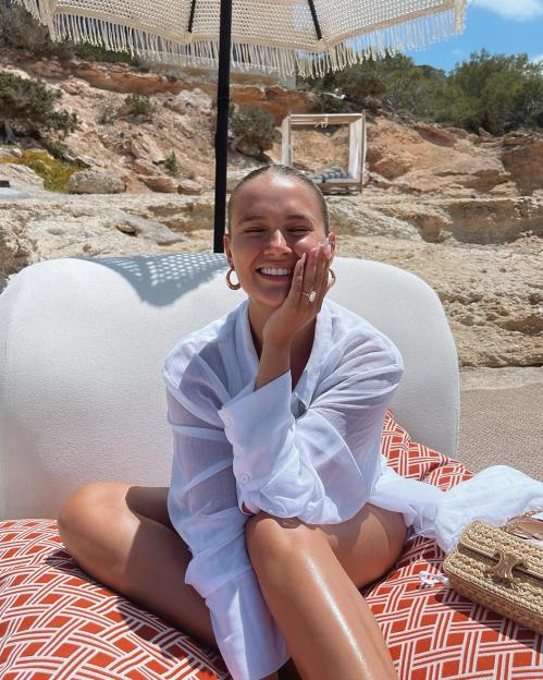 Molly Mae sitting under a white fringed parasol, smiling and showing off her engagement ring.