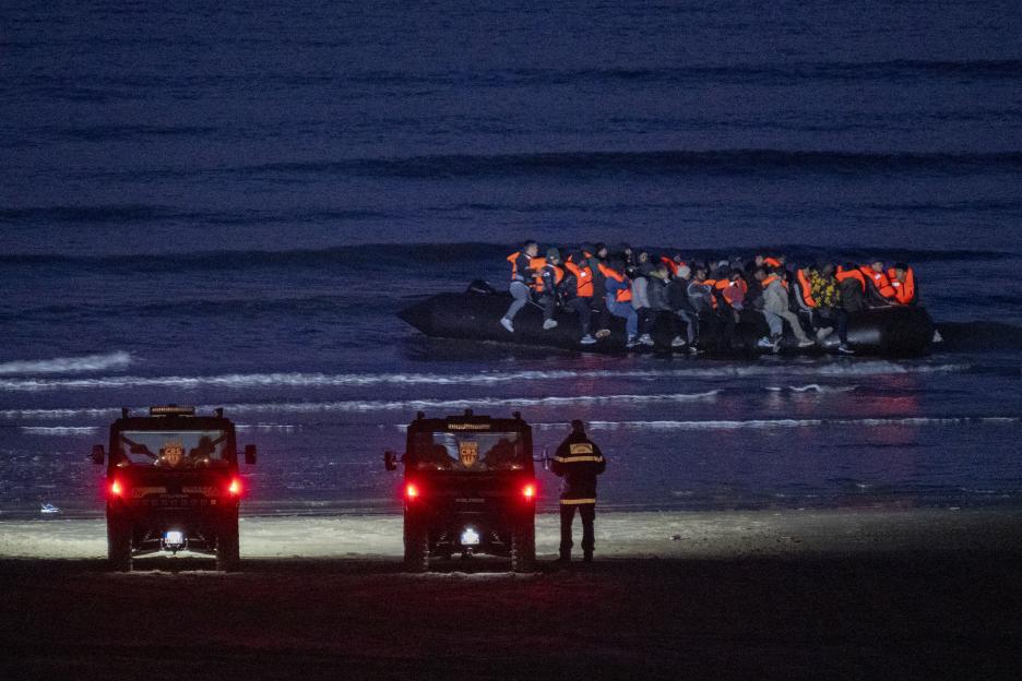 A group of migrants in life jackets in an inflatable boat on the water as police in two vehicles and one officer stand on the beach.