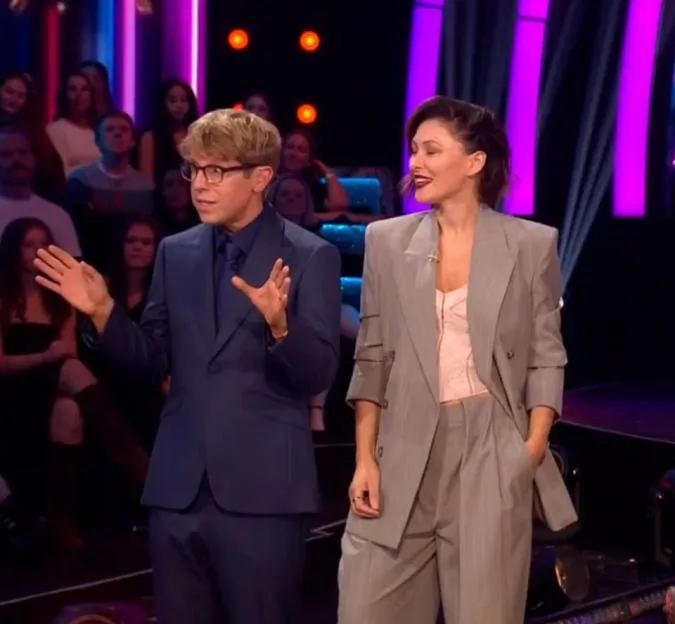 Two TV show hosts, a man in a blue suit talking with hand gestures and a woman in a grey pinstripe suit, standing on stage in front of an audience and purple lighting.