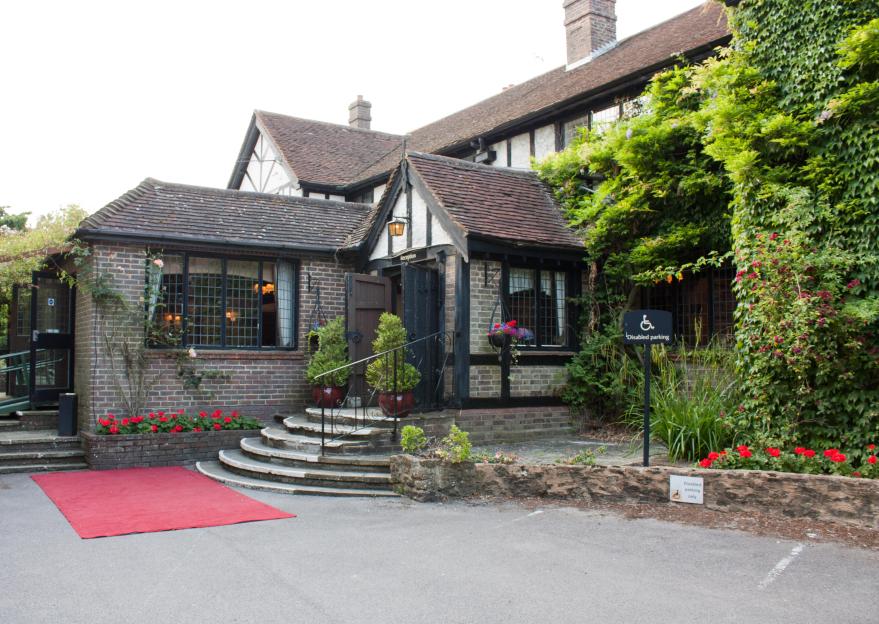 Cisswood House, a building with brickwork, tiled roofs, and lush greenery, featuring a red carpet leading to an entrance with steps and a "Reception" sign, and a "Disabled parking only" sign.