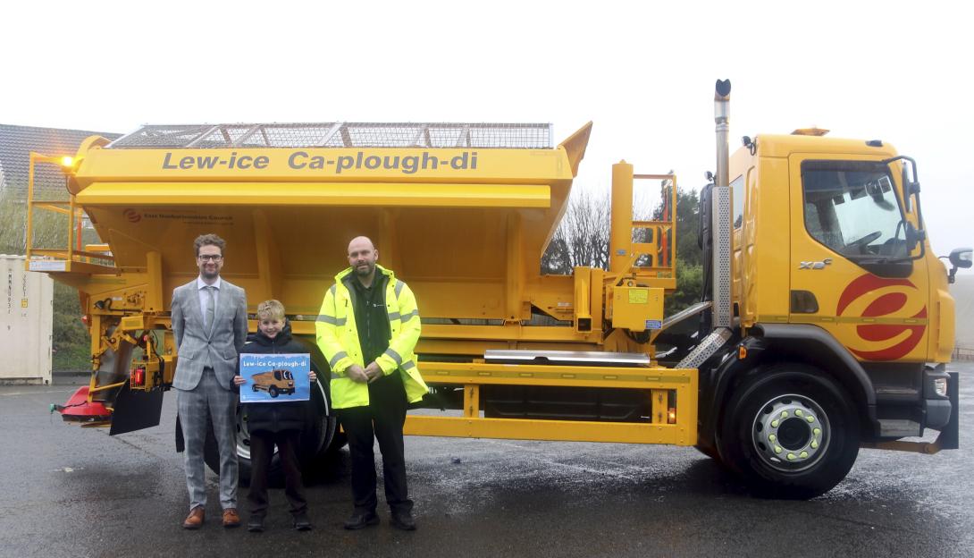 Three individuals stand in front of a yellow gritter truck with "Lew-ice Ca-plough-di" written on its side.