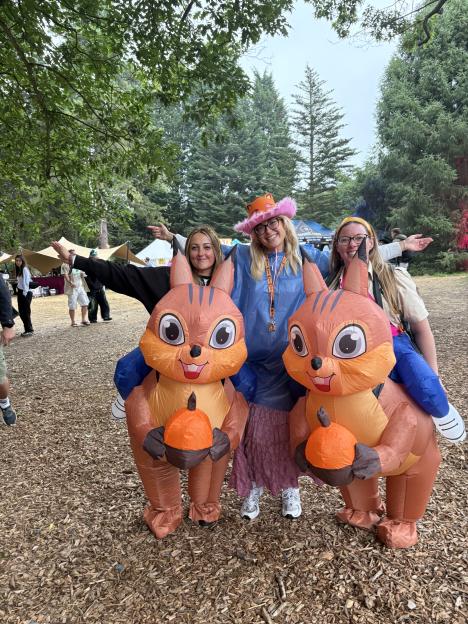 Three women pose in a park, two wearing inflatable squirrel costumes and one in a pink fuzzy hat and blue poncho.