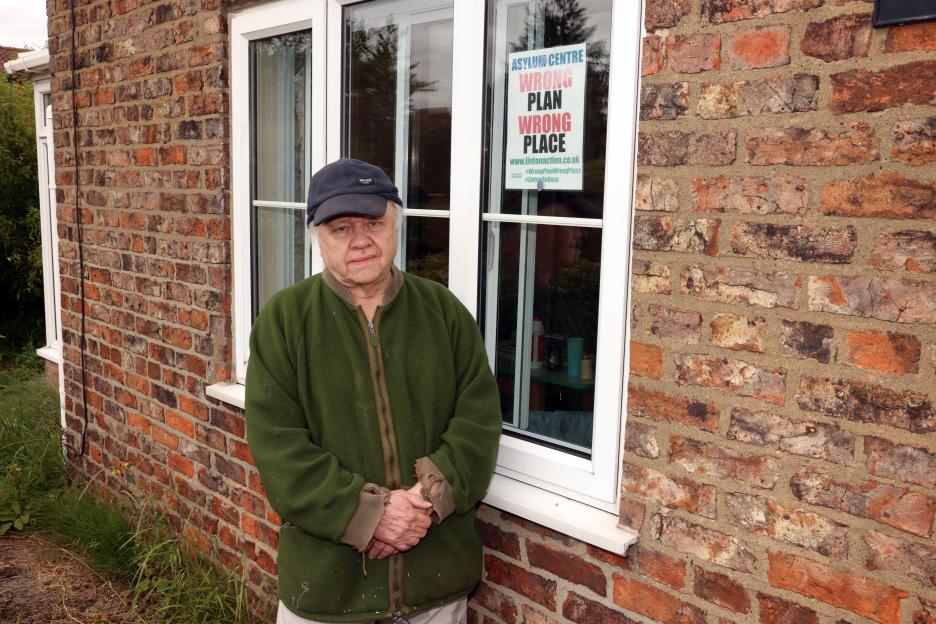 Local resident Trevor Dawson stands in front of a brick house with an "Asylum Centre Wrong Plan Wrong Place" sign in the window, expressing local outrage about a planned asylum seeker center in RAF Linton On House.