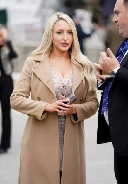 A blonde woman in a tan coat and houndstooth dress holds a phone while talking to a man in a suit at the Cheltenham Festival 2024.