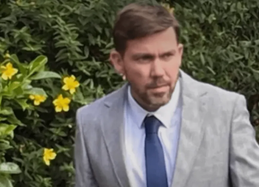 A man in a suit and tie, against a backdrop of green foliage with yellow flowers.