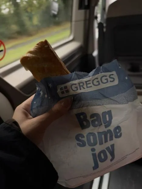 A person holding a Greggs pastry in a paper bag inside a vehicle.