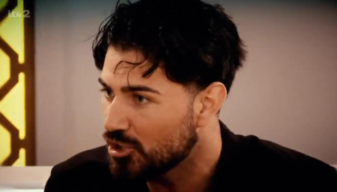 A man with dark hair and a beard, in a dark shirt, looks off to the side, mouth slightly open.