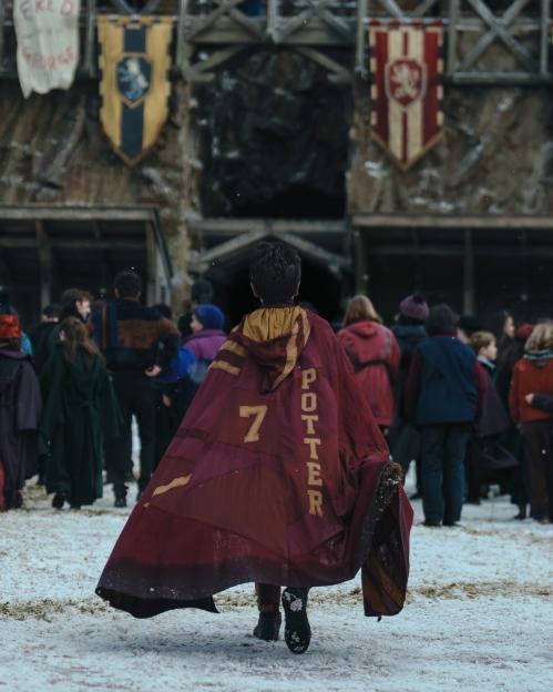 A child in a cape with "POTTER" and "7" on the back walks in the snow toward a building with house banners.