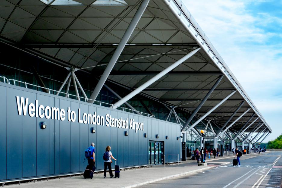 Passengers with luggage walking towards the entrance of London Stansted Airport with the welcome sign.