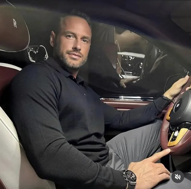 A man in a black polo shirt and grey trousers sitting in the driver's seat of a luxury car, holding the steering wheel and looking at the camera.