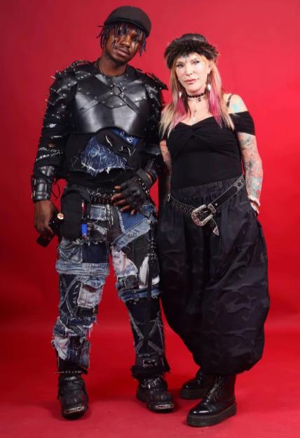 A man and a woman in punk-style clothing standing against a red background.