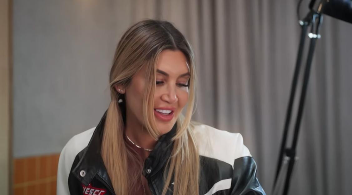A smiling woman with blonde hair, wearing a black and white leather jacket, looks down and to the right.