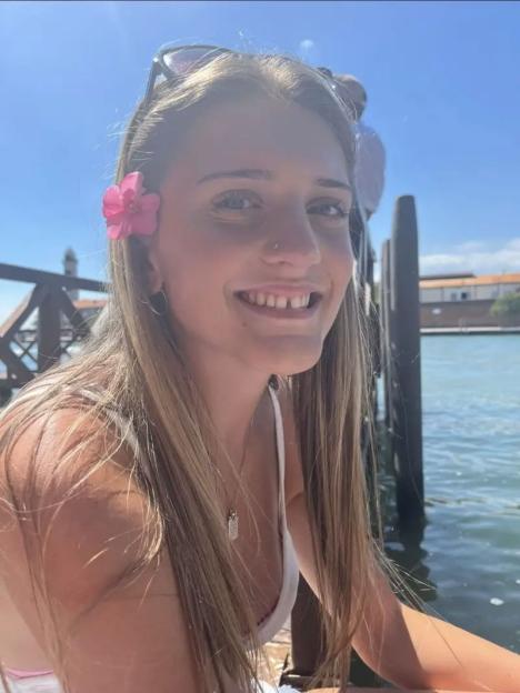 A young woman with long blonde hair and a pink flower in her hair smiles while sitting by the water on a sunny day.
