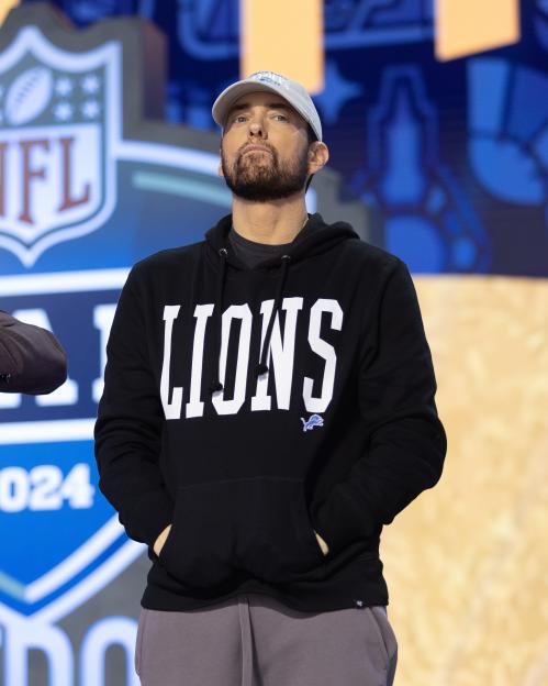 Eminem wearing a black Detroit Lions hoodie and a white baseball cap, looking up.