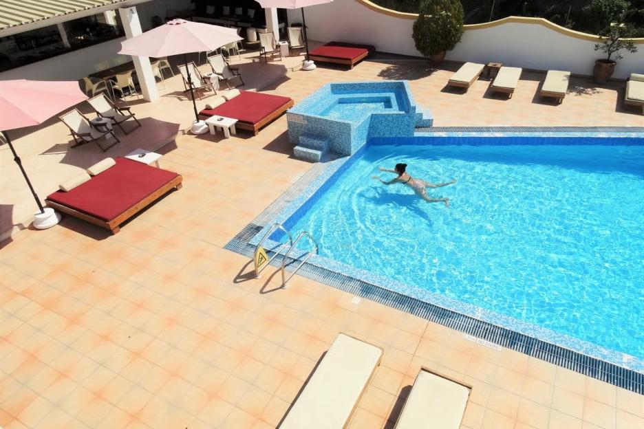 Overhead view of a woman swimming in a bright blue pool surrounded by sunbeds and umbrellas.