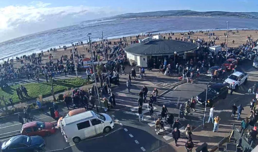 Large crowd gathered at Budleigh Salterton beach, where vehicles are lined up along the street.