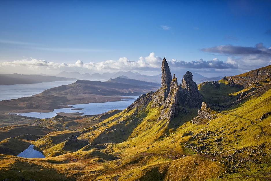 Old Man of Storr rock formations on the Isle of Skye, Scotland, with a loch and mountains in the background under a blue sky.