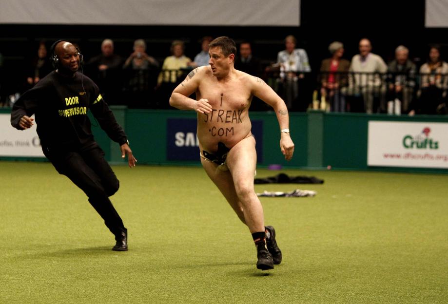 A streaker runs across a green floor while a security guard chases him.