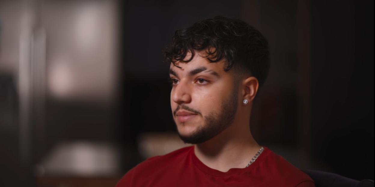 A man with dark curly hair, a beard, and a silver chain on his neck is looking to the right.
