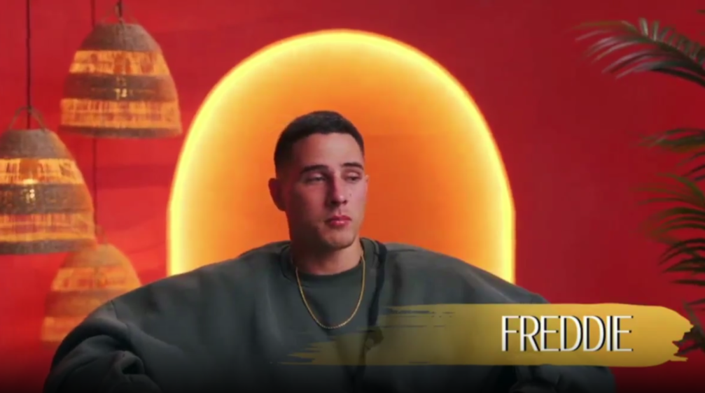 Freddie in a gray sweatshirt with a gold chain, set against a red background with a large orange circle and hanging woven lamps.
