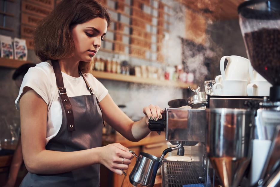 A young female barista steams milk at a coffee machine.