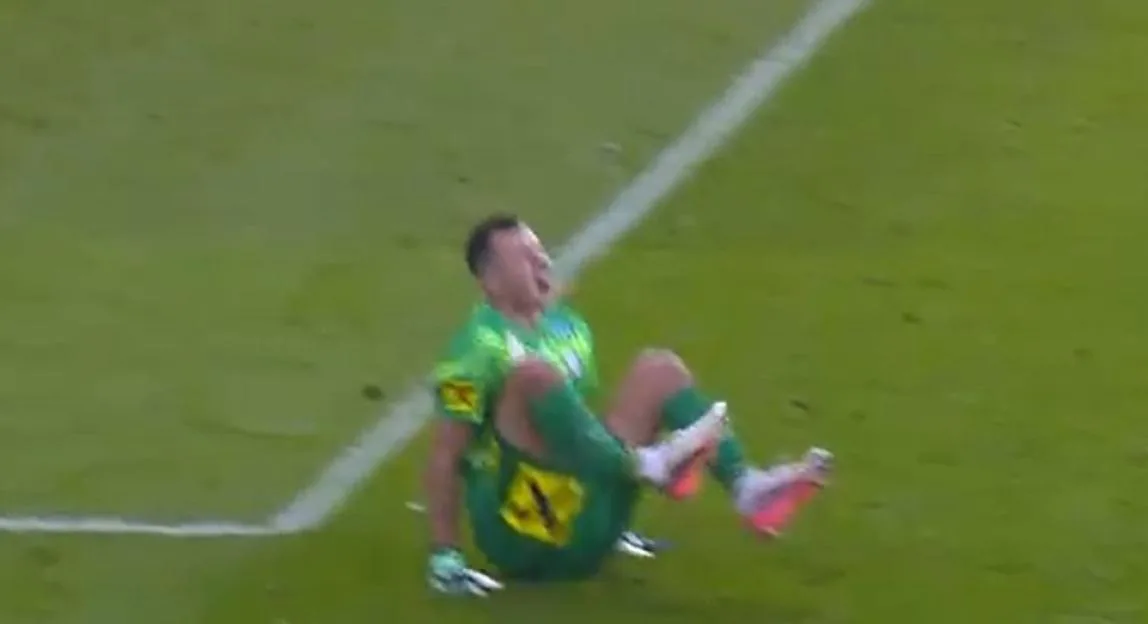 A goalkeeper sitting on the grass in pain after suffering a leg injury.