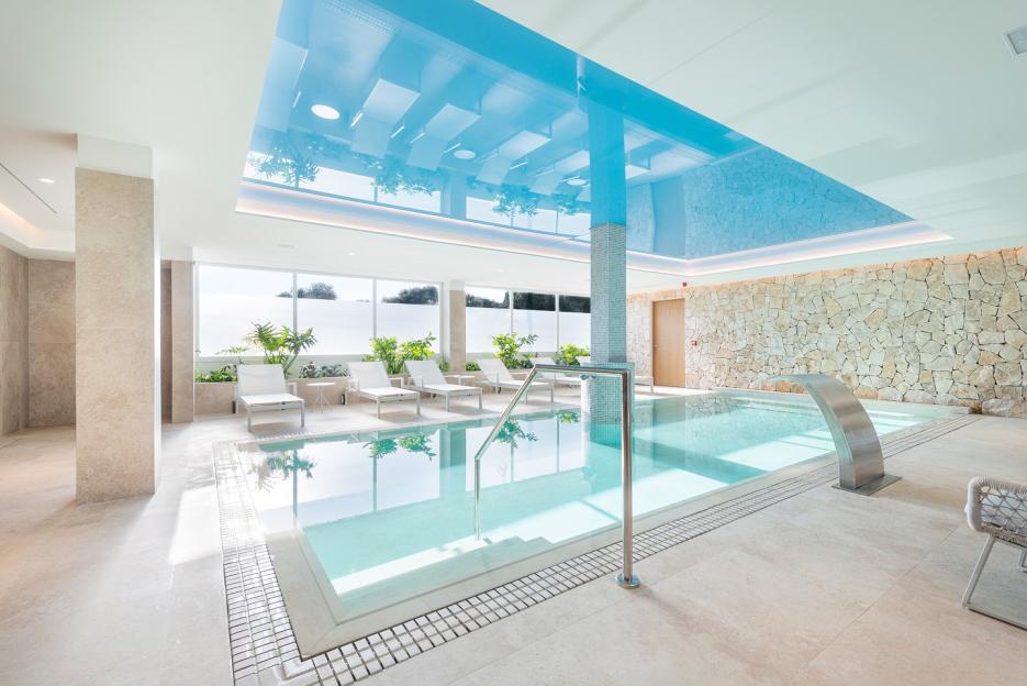 An indoor hotel pool with white lounge chairs, light stone walls, and a light blue ceiling.