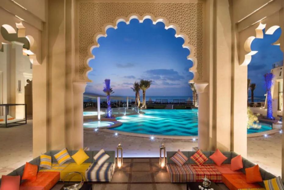 A luxurious resort with a swimming pool and ocean view, framed by an ornate archway and lounge seating.