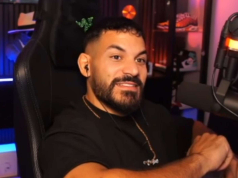 A man with a beard and short dark hair sits in a gaming chair, looking to his right.