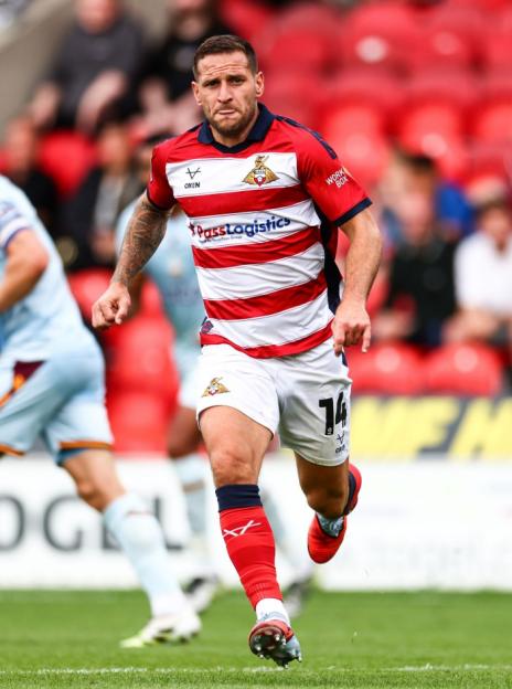 A male football player in a red and white striped jersey, white shorts, and red socks runs on a green field.